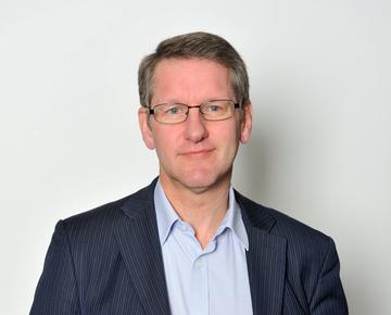 A white man with blonde hair and blue eyes smiles at the camera. He wears glasses and a blue shirt and striped blazer.