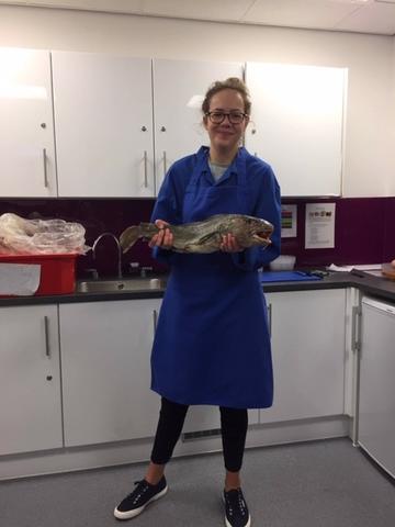 A blond-haired white woman stands in a blue coat, holding a fish. Behind her are white kitchen cabinets and a red bucket on the counter.