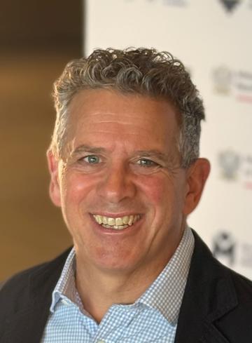 A white man with short grey curly hair and blue eyes smiles at the camera. The top of his shirt collar (blue check) and blazer are visible.
