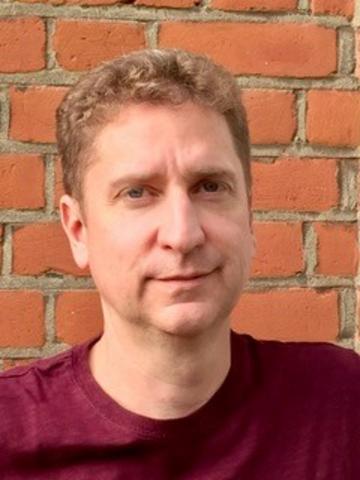 A white man with short curly blonde hair smiles at the camera. He is wearing a maroon t-shirt.