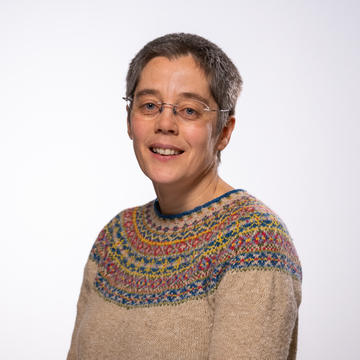 A white woman with short dark hair smiles at the camera. She is wearing a Fair Isle jumper.