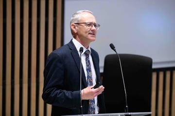 A white man with grey hair and glasses is speaking at a podium. He is wearing a blue blazer, white shirt and floral tie.