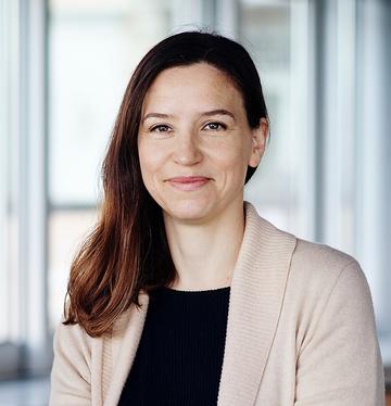 A white woman with long dark hair smiles at the camera. She wears a cream cardigan over a black shirt.