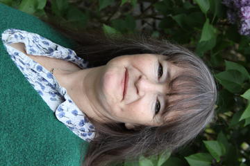 A white woman with dark brown-grey hair looks into the camera. There are green leaves in the background and she is wearing a green jersey and collared shirt.