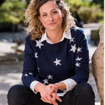 A white woman with curly brown hair smiles at the camera. She is wearing a dark blue jersey with white stars, and a white collared shirt underneath.