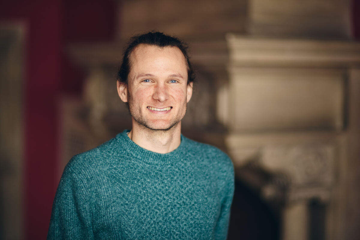 A white man with brown hair pulled back smiles at the camera. He is wearing a seagreen jumper.