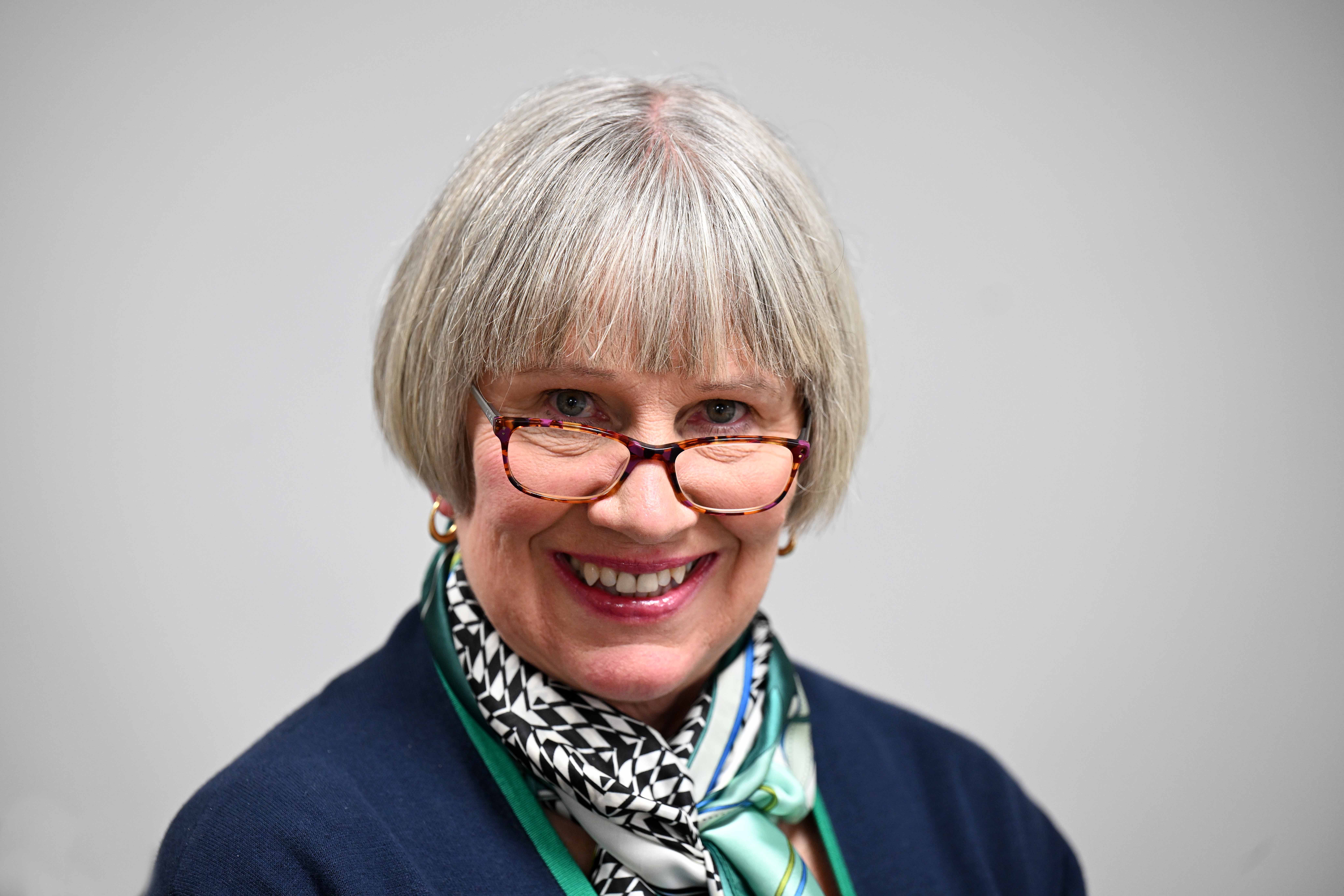 A white woman with grey hair cut into a bob smiles at the camera. She wears glasses and has on a navy blue blazer