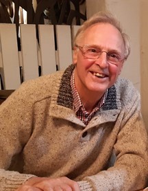 A white man in a cream collared jumper smiles at the camera. He has blonde hair and glasses.