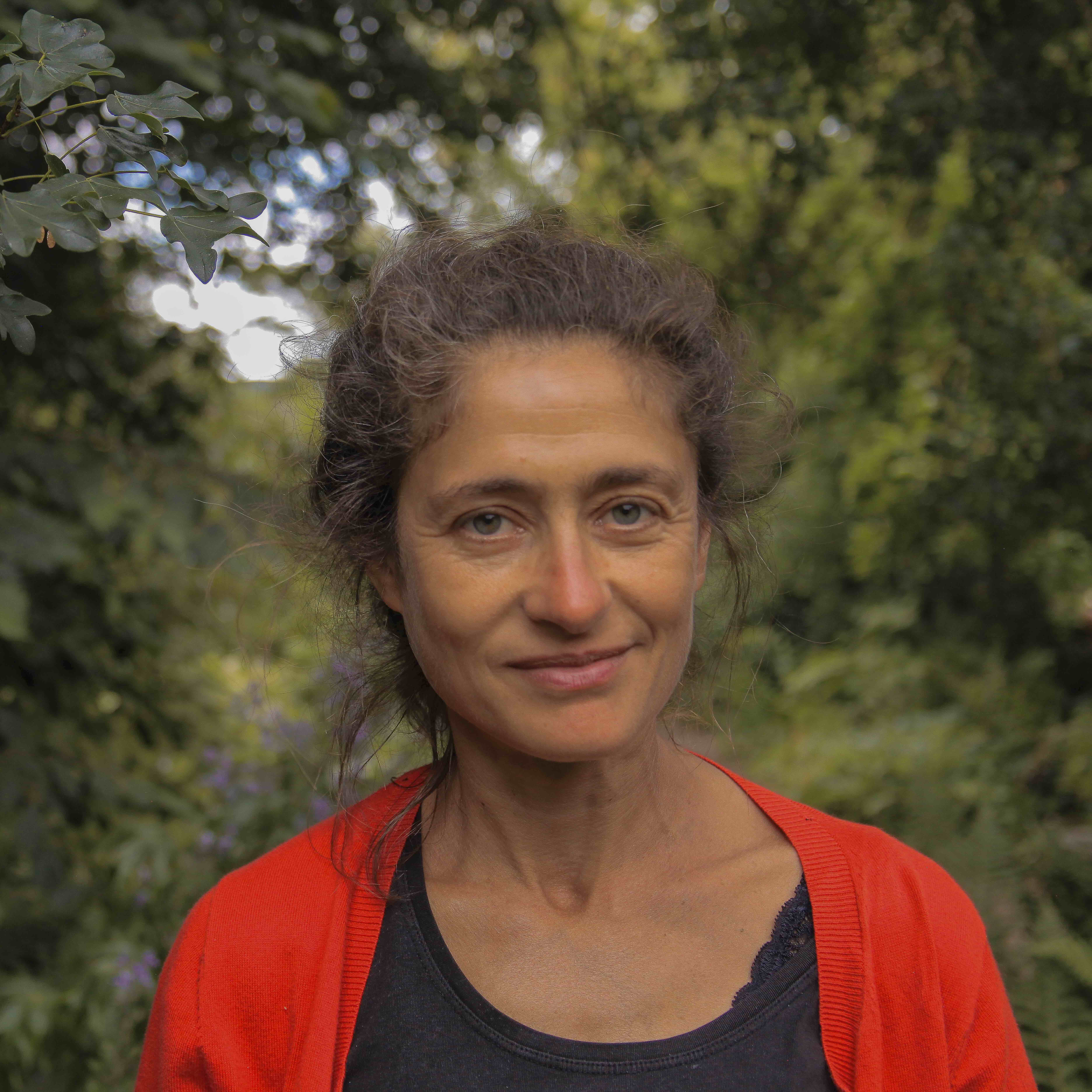A white woman smiles at the camera. She is surrounded by green leaves and wears a bright red cardigan over a black t-shirt