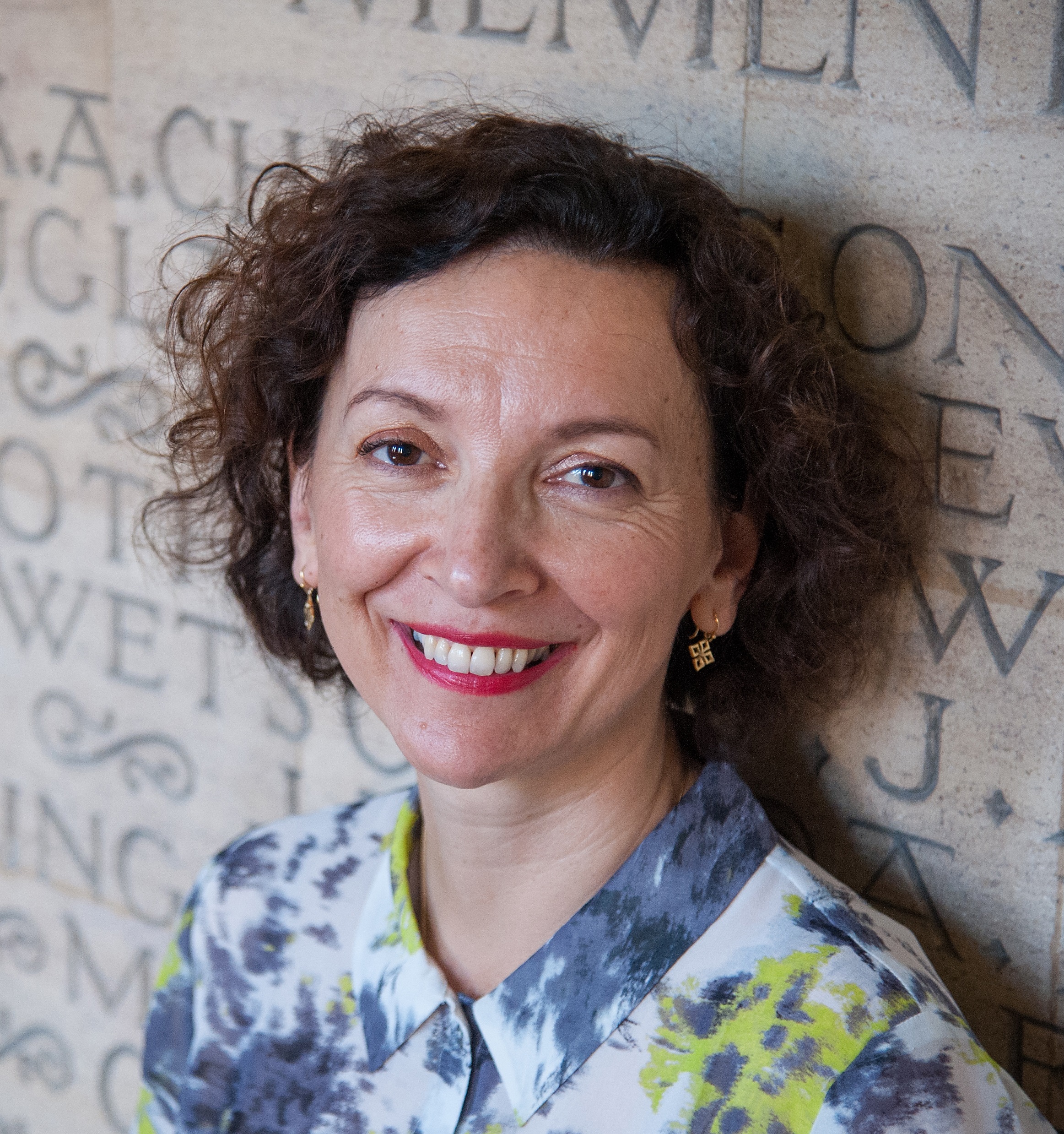 A white woman with brown curly hair smiles at the camera. She wears a blue and yellow-green shirt.