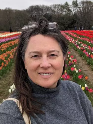 A white woman with dark hair and a grey poloneck jumper on stands in a field of tulips. She has glasses perched on her head and is smiling at the camera.