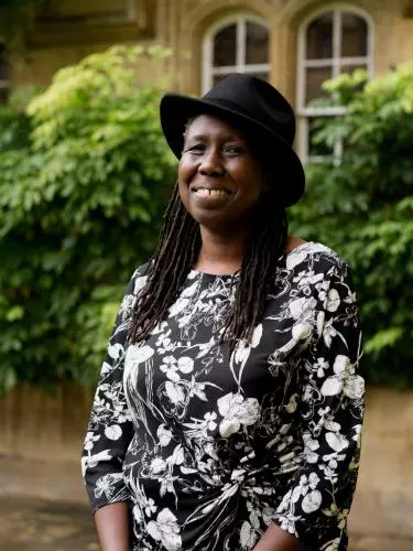 A black woman with long braids smiles at the camera. She wears a black hat and a black and white patterned dress.
