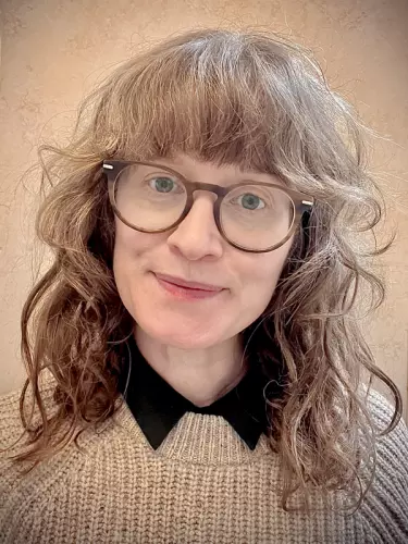 A white woman with dark curly hair and blue eyes smiles toward the camera. She is wearing glasses and a beige fisherman jersey.