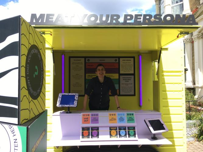 A bright yellow horsebox centres the picture with 'Meat Your Persona' written across the top. Inside a white woman with short dark hair is smiling. She stands behind lilac shelves which hold postcards. There are two tablets at either end of the horsebox.