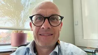 A bald white man wearing glasses smiles at the camera. He has a blue shirt on and a plant in the background.