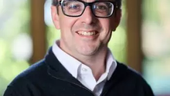 A white man with glasses on smiles at the camera. He has dark hair and wears a dark jumper over a pink shirt
