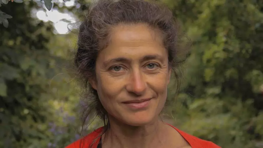 A white woman smiles at the camera. She is surrounded by green leaves and wears a bright red cardigan over a black t-shirt