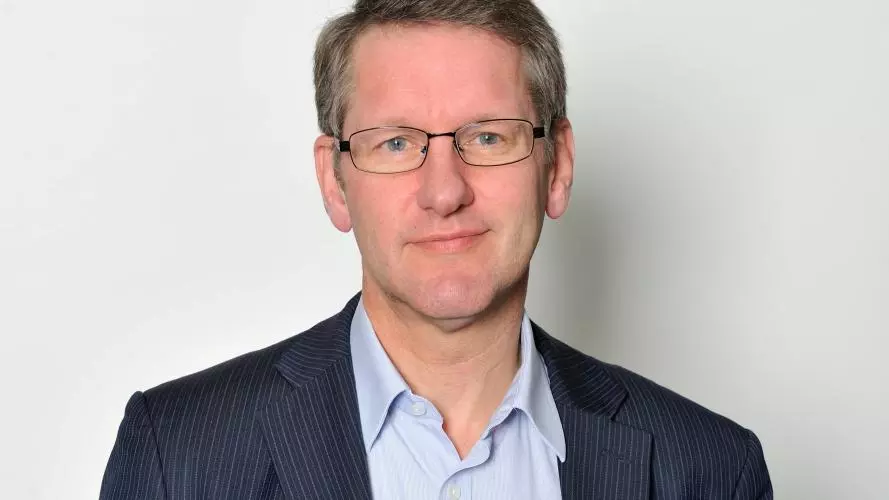 A white man with blonde hair and blue eyes smiles at the camera. He wears glasses and a blue shirt and striped blazer.
