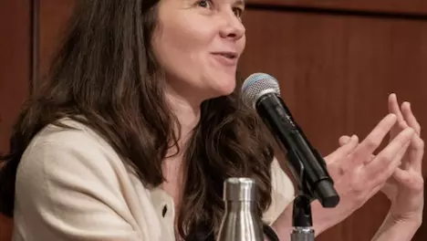 A white woman with dark hair and wearing a cream cardigan, talks with her hands. She has a microphone in front of her.