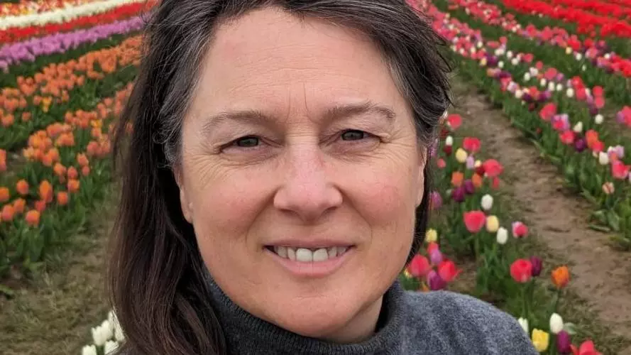 A white woman with dark hair and a grey poloneck jumper on stands in a field of tulips. She has glasses perched on her head and is smiling at the camera.