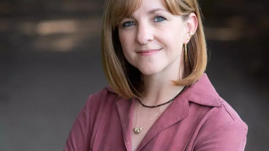 A red haired white woman smiles at the camera. She is wearing a magenta top and has her arms crossed.