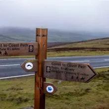 A sign showing the directions for the Wainwright Way, including the distinctive 'AW' initials at the top of the sign. Behind the sign is road and then rolling hills shrouded in mist.