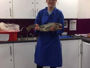 A blond-haired white woman stands in a blue coat, holding a fish. Behind her are white kitchen cabinets and a red bucket on the counter.