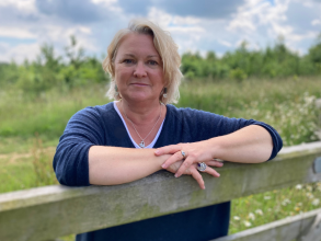 A white woman leans over a farm gate. She has bobbed blonde hair and wears a dark blue v-neck jersey. Behind her is a green field and blue sky with clouds.