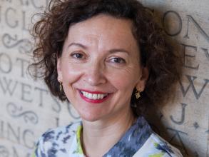A white woman with brown curly hair smiles at the camera. She wears a blue and yellow-green shirt.