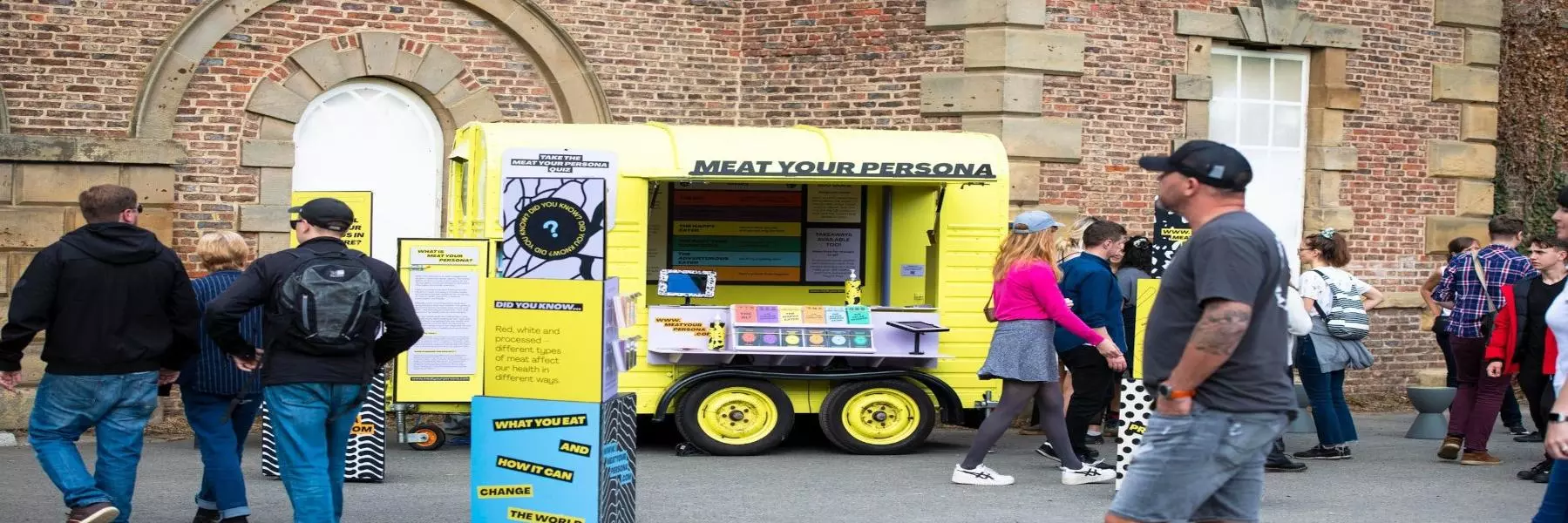 A bright yellow horsebox sits in front of a redbrick building. The horsebox reads Meat your Persona and has a pop up display in front of it. People are walking past.