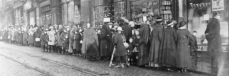 A black and white photograph from world war one showing people queuing in the street, waiting for shops to open